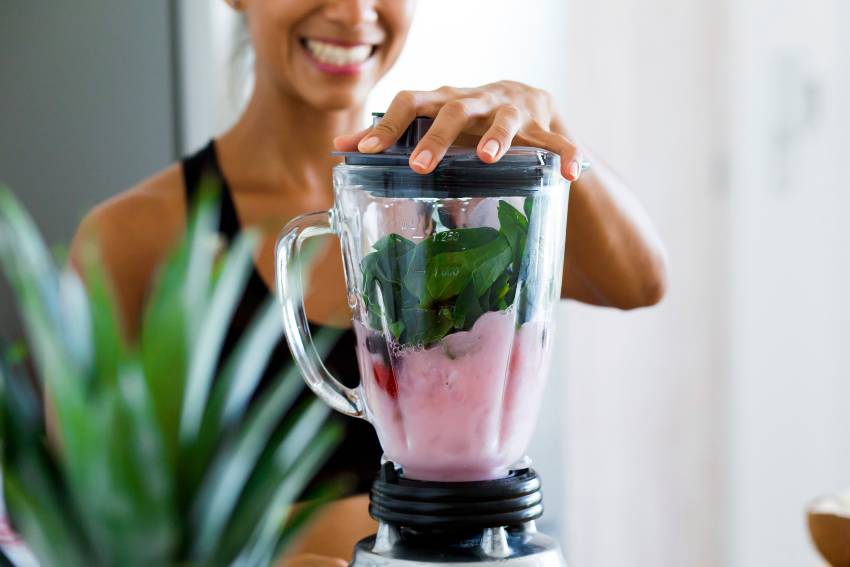 A woman making a green and pink smoothie with a blender 